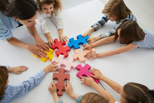 Team Of Elementary School Children Together With Teacher Join Colorful Jigsaw Puzzle Pieces Standing Around White Table In Classroom, Top View Close Up. Education, Teamwork, And Fitting In With Group