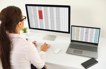 Young woman sitting at white office desk, looking at screens of desktop and laptop computers, working with data lists, electronic business documents and spreadsheets. Back view, over shoulder