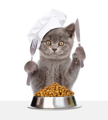 Poster - Kitten wearing chef's hat eats dry food with fork and knife from bowl. isolated on white background