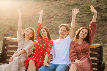 company of beautiful young people in a summer park on a bench with raised hands 