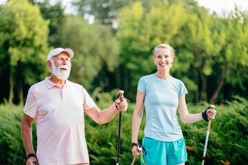 Portrait of senior man and adult woman enjoying a nordic walk. Active elderly man and middle age blond sporty woman walking on the park and smiling.