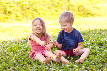 A blonde-haired boy and a girl are sitting on a green grass lawn and playing with each other. Happy childhood, fun vacation