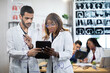 © sofiko14 - Two multiethnic male and female doctors standing behind glass wall with MRI scan and working on tablet pc. Their colleagues talking each other at the table on background