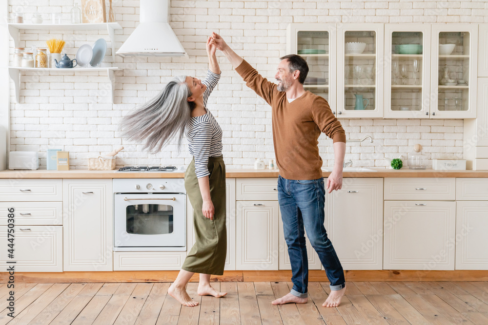 Mature family middle-aged couple wife and husband dancing together in the kitchen, celebrating date anniversary. Active seniors, love and relationship concept. Social distance. - obrazy, fototapety, plakaty 