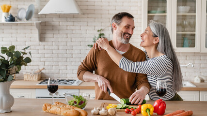 Cheerful mature husband man cutting vegetables cooking food meal in the kitchen while his wife woman embracing hugging him helping prepare salad together at home