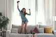 © nenetus - Beautiful woman listening to music with headphones and dancing on the couch in the living room at home.