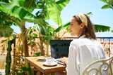 Fototapeta  - Young beautiful mixed race woman sitting on terrace at morning in hotel, using laptop computer while enjoying vacation