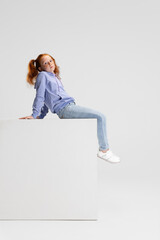 Wall Mural - One cute red-headed girl in casual clothes sitting on big box isolated on white studio background. Happy childhood concept. Sunny child. Looks happy, delighted