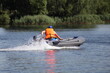One man in orange lifejacket fast floats on inflatable motor boat with small outboard motor on the river on green grass background on shore, active recreation on the water at Sunnny summer day
