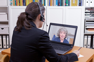 Female lawyer with headset in front of her laptop writing something on a paper while having a live video chat with an elderly client, copy space