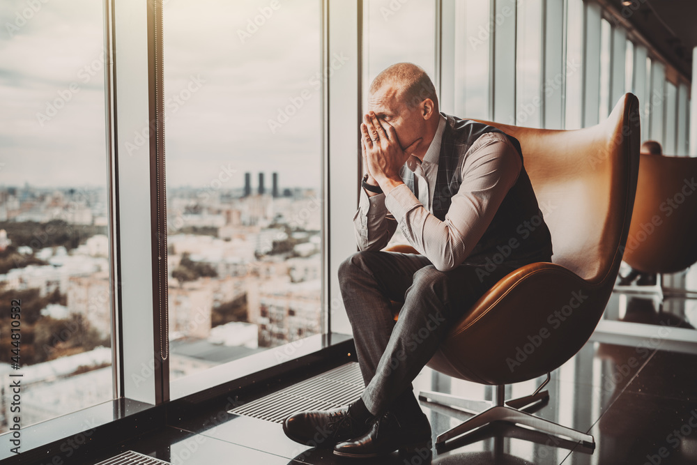 A stressed out adult caucasian man entrepreneur partly closing face with his hands while sitting on an office armchair next to the window of a modern business skyscraper and looking on the cityscape - obrazy, fototapety, plakaty 