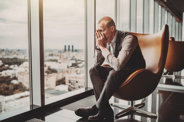 Wall Mural - A stressed out adult caucasian man entrepreneur partly closing face with his hands while sitting on an office armchair next to the window of a modern business skyscraper and looking on the cityscape