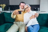 Fototapeta Londyn - Happy mature senior couple taking selfie looking at smartphone, cheerful elderly grandparents having fun holding phone, taking self portrait picture laughing sitting on sofa