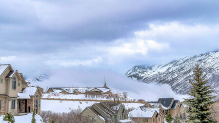 Wall Mural - Pano Snow covered mountainscape with houses on the slopes under cloudy sky in winter