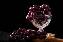 Red, Black Grapes On A Brown Wooden Cutting Board. Dark, Black Background. Morning Light, Ray Shining On The Grapes And Crystal. Retro, Bohemic Style.