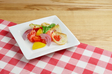 Fresh tomatoes salad with onion and croutons served in a square plate over rustic wooden table with red plaid tablecloth