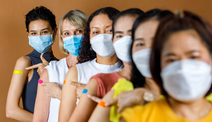 Diverse group of multinational ethnic female patients wears face mask stand in line by height order look at camera pointing colorful plasters on shoulder together after vaccinated