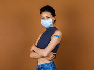 Wall Mural - Portrait Closeup studio shot of cute confident beautiful sexy female patient wears face mask stand crossed arms show blue plaster on her shoulder after receive Coronavirus Covid-19 vaccine injection