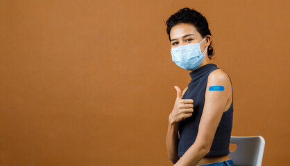 Wall Mural - Happy beautiful sexy female patient wears face mask stand show thumb up at blue plaster on her shoulder after receiving Coronavirus Covid-19 vaccine injection