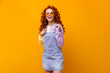 Joyful woman with curly red hair looks into camera. Girl in sunglasses, sundress and sweatshirt is smiling on orange background