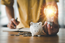 Saving Energy And Money Concept. Idea For Save Or Investment. Businessman Holding Lightbulb Beside Piggy Bank And Coins Stacking On Desk With Note Book.