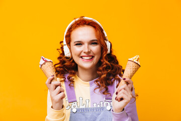 Wall Mural - Positive girl with curls laughs, looks into camera and holds ice cream. Woman in headphones posing on orange background