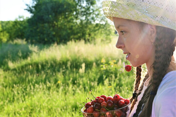 in summer garden young sweet girl in white T-shirt and hat holds  ripe cherry berry in her mouth. Delicious and healthy food. Close-up