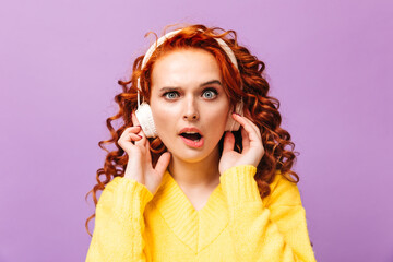 Wall Mural - Girl looks at camera shocked against purple background. Redhead woman listening to music on headphones