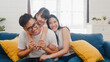 © tirachard - Happy cheerful Asian family dad, mom and daughter having fun cuddling and video call on laptop on sofa at house. Self-isolation, stay at home, social distancing, quarantine for coronavirus prevention.