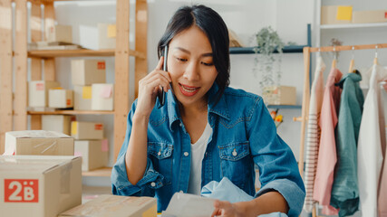 Wall Mural - Young Asia businesswoman using smartphone call receiving purchase order and check product on stock work at home office. Small business owner, online market delivery, lifestyle freelance concept.