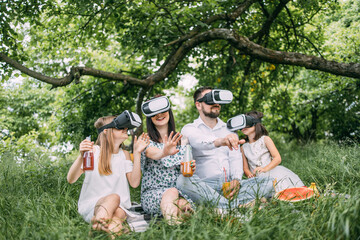 Parents and their two daughters wearing VR headset during picnic at summer garden. Young family having fun with innovative gadgets outdoors. Modern lifestyles.