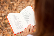 Woman Reading Poems In Autumn Park. Back View Of Opened Book With Red Cover In Hands Of A Girl.