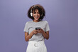 © Look! - Joyful girl in headphones holding smartphone on purple background. Happy curly woman in gray tee listening to music on isolated backdrop