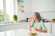 Portrait of attractive elderly dreamy grey-haired woman drinking coffee eating yummy tasty bakery at home light white indoors