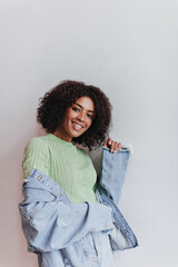 Wall Mural - Dark-skinned girl in green top takes off her jeans jacket. Portrait of attractive woman in denim jacket and skirt smiling on white backdrop