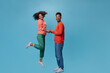 © Look! - Couple holds hands and smiles on blue background. Curly brunette woman in green pants jumps and dark-skinned man looks into camera