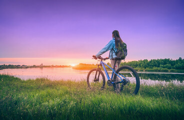 Wall Mural - woman with bicycle near the river at sunset in spring. landscape with sporty girl with backpack ridi