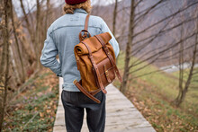 Young Man Exploring Nature Landscapes In Spring, Walking Across Alone With Brown Backapack. Handsome Hiker Traveling In Forests. Mountains In The Background, Rear View, Focus On Leather Backpack