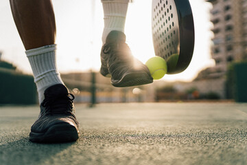 Man lifting paddle ball with his racket and shoe on the green court during training in the sunset