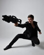 Full length portrait of a  brunette man wearing leather jacket  and holding a science fiction gun.  kneeling  action pose isolated  against a grey studio background.
