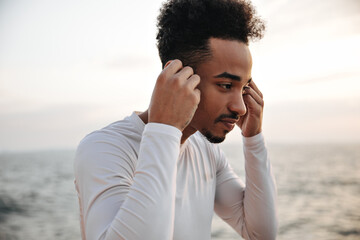 Wall Mural - Beautiful man in white long-sleeved t-skirt puts on wireless headphones. Portrait of dark-skinned sportsman near sea.