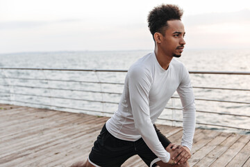 Wall Mural - Handsome curly brunette man in black shorts and long-sleeved t-shirt stretches outside. Dark-skinned guy works out near sea.