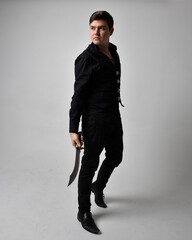 Wall Mural - Full length portrait of a  brunette man wearing black shirt and gothic waistcoat holding sword.  Standing  action pose isolated  against a grey studio background.
