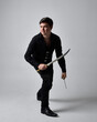 Full length portrait of a  brunette man wearing black shirt and gothic waistcoat holding sword.  Standing  action pose isolated  against a grey studio background.
