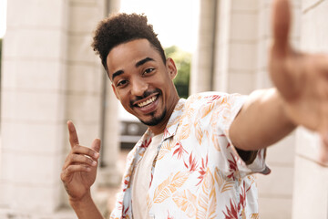 Wall Mural - Excited bearded charming man in white floral summer shirt points up, looks into camera, smiles and takes selfie outside.