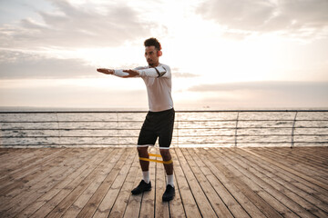 Wall Mural - Full-length portrait of brunette curly man in black shorts and white long-sleeved t-shirt listening to music and squatting near sea.