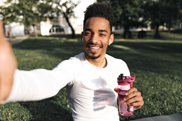 Wall Mural - Attractive young curly dark-skinned man in white sport t-shirt smiles sincerely, holds pink water bottle and takes selfie in park.