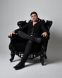Full length portrait of a  brunette man wearing black shirt and  waistcoat.  Seated  pose in a gothic throne chair isolated  against a grey studio background.