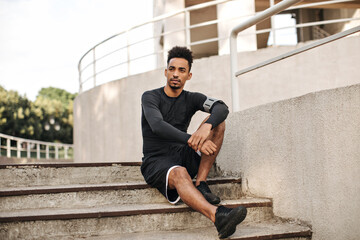 Wall Mural - Brown-eyed brunet dark-skinned bearded man in long-sleeved t-shirt and shorts sits on stairs outside and looks away.