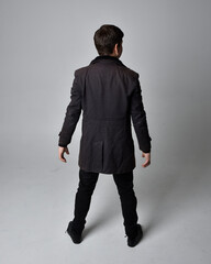 Wall Mural - Full length portrait of a brunette man wearing black leather coat.  Standing pose facing backwards, walking away from the camera isolated  against a grey studio background.